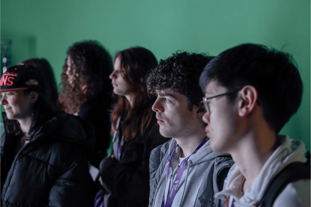 Six young people stood in front of a green screen watching something off camera.