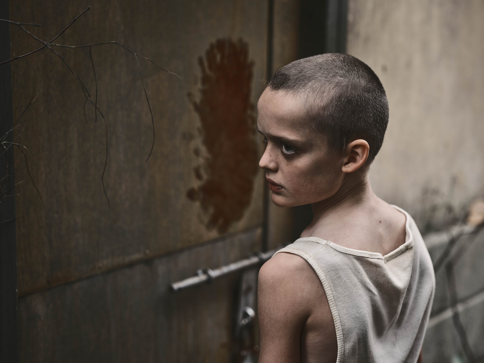 A child with a shaved head and vest stares ominously.