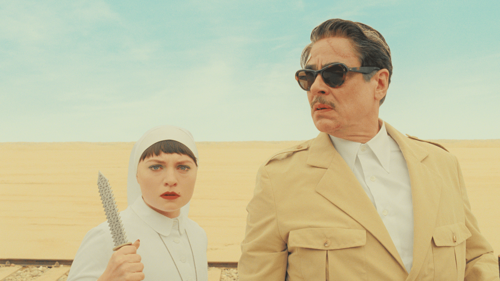 A man in a safari jacket stands beside a nun holding a knife in front of a sand dune.