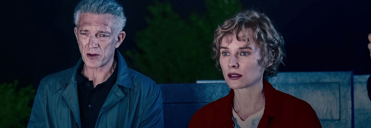 A man and woman stare off camera, seemingly disturbed. He has grey hair and a jack while she wears a black overcoat.