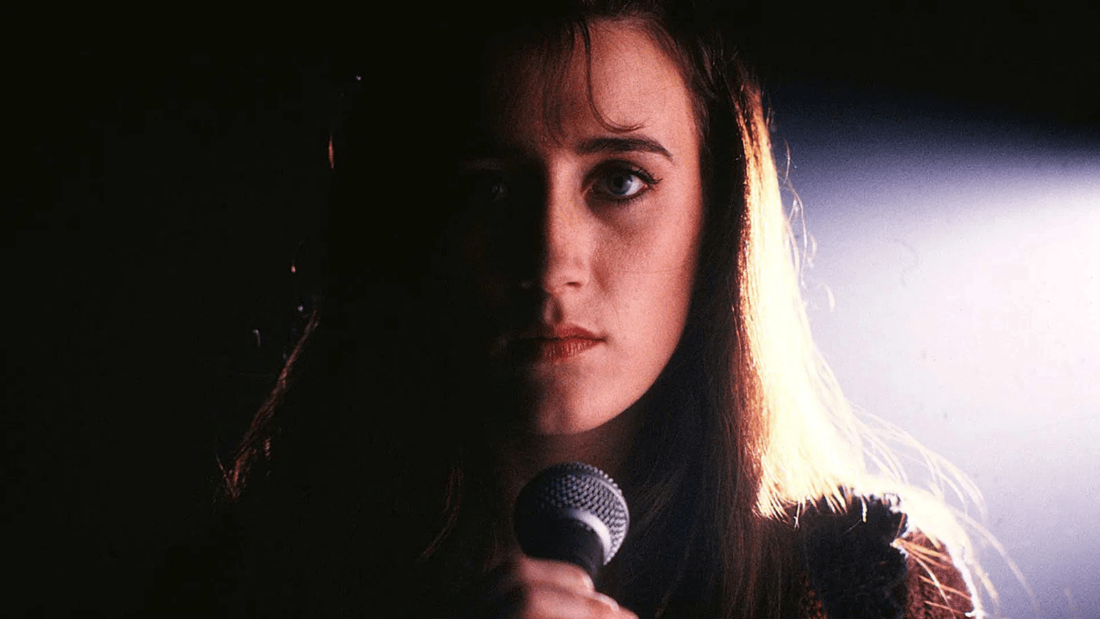 A young woman stands on stage in dramatic half-light, holding a microphone and staring solemnly forward, her face partially obscured by shadow.